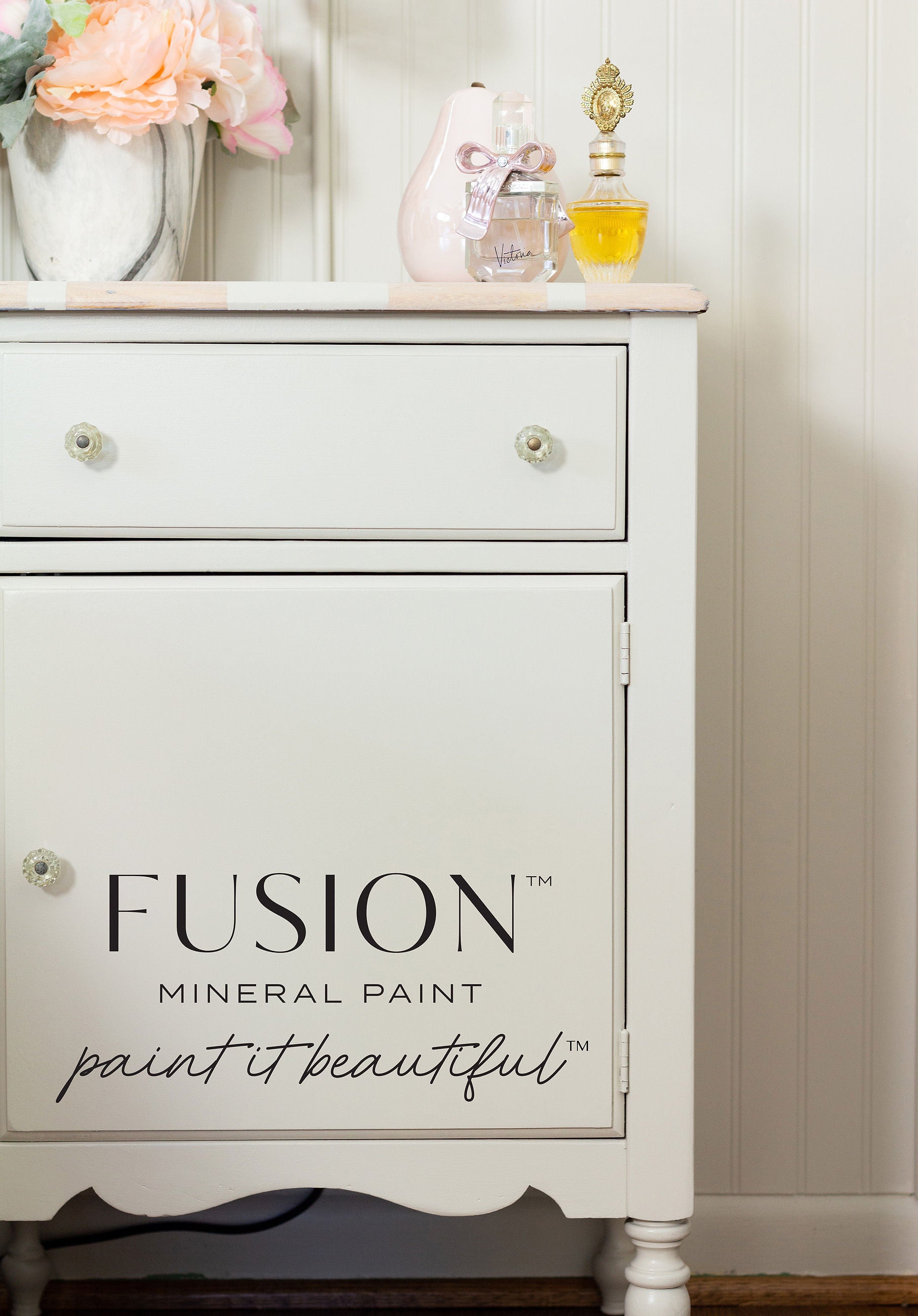 Night stand painted in color chateau with perfume bottles on top with a vase of flowers in colors pink and orange. 